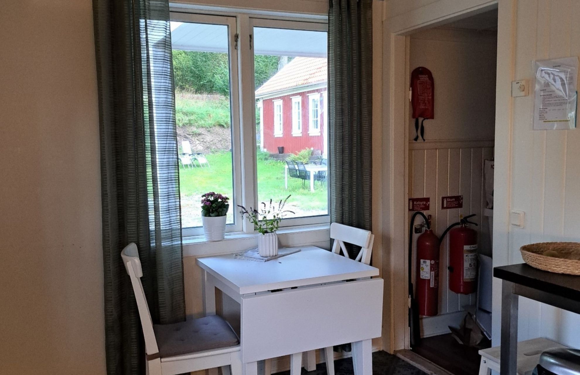 Litet köksbord med två stolar vid ett fönster med utsikt över en trädgård och röd byggnad