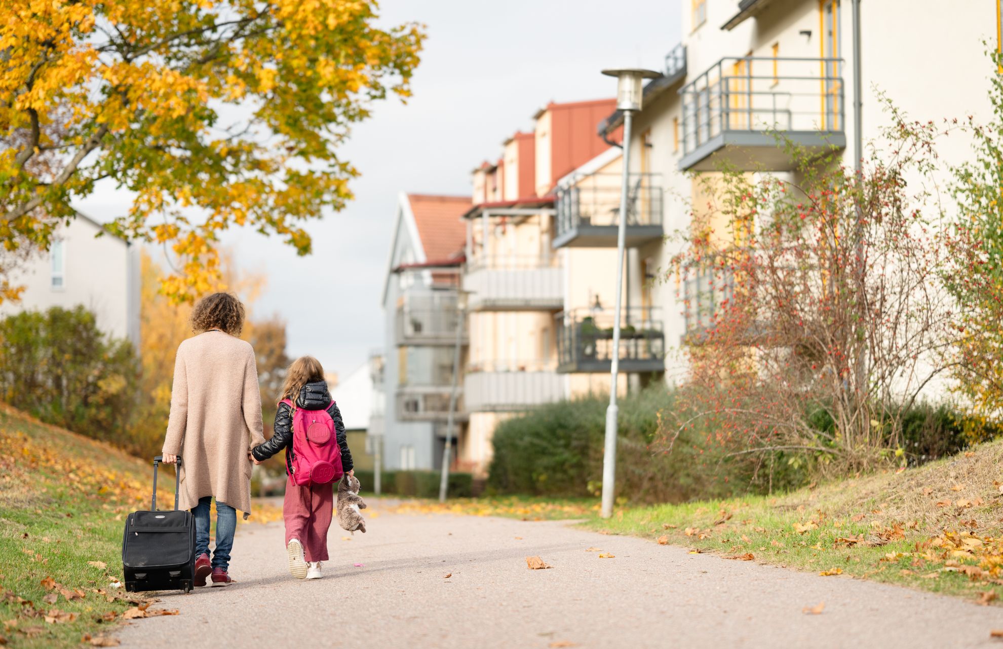 Vuxen och barn promenerar längs bostadsgata i höstmiljö med rullväska och ryggsäck