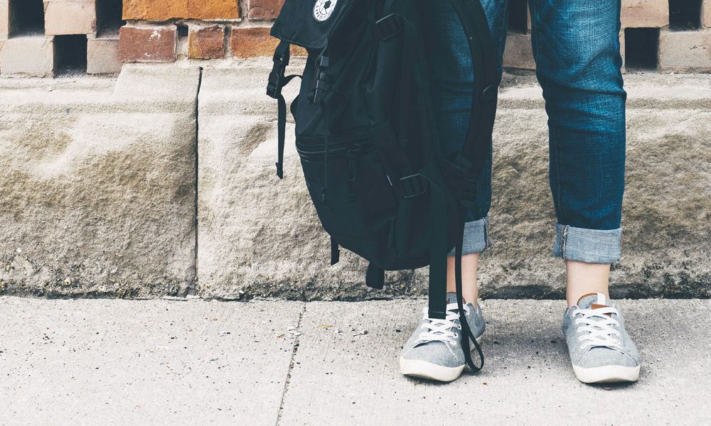 Ungdom med jeans och gråa sneakers håller en svart skolryggsäck framför en tegelvägg
