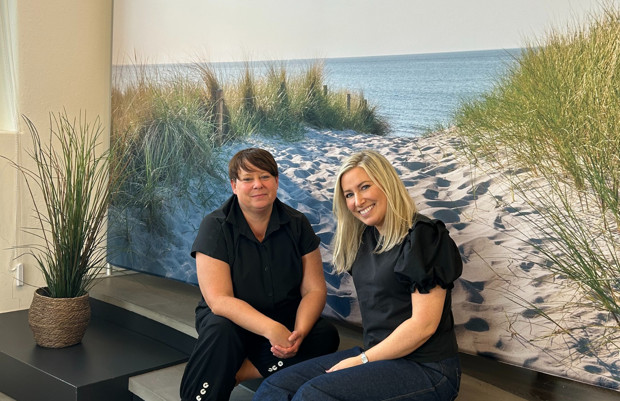 Viljans metodteam sitter framför en strandmålning med gräs och hav, leende och klädda i svarta kläder