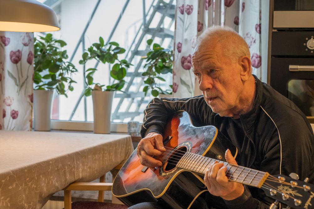 Äldre man spelar gitarr i kök, omgiven av blommiga gardiner och krukväxter