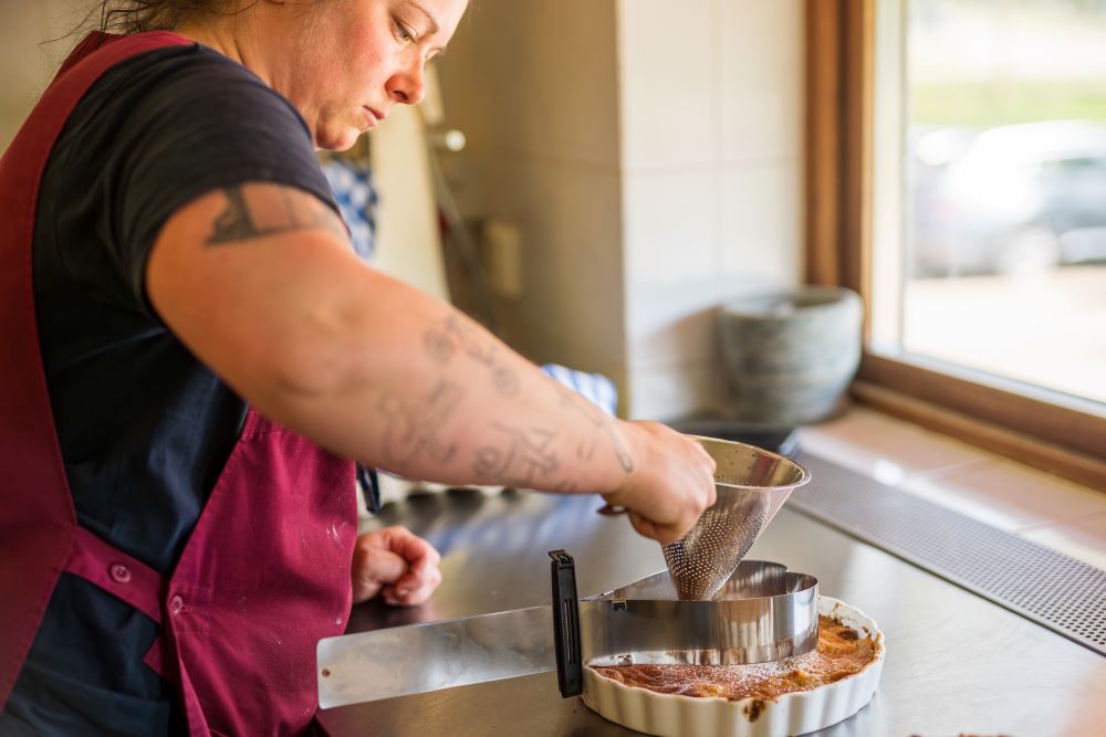 En person i ett kök som förbereder en paj med hjälp av en sil för att tillsätta florsocker