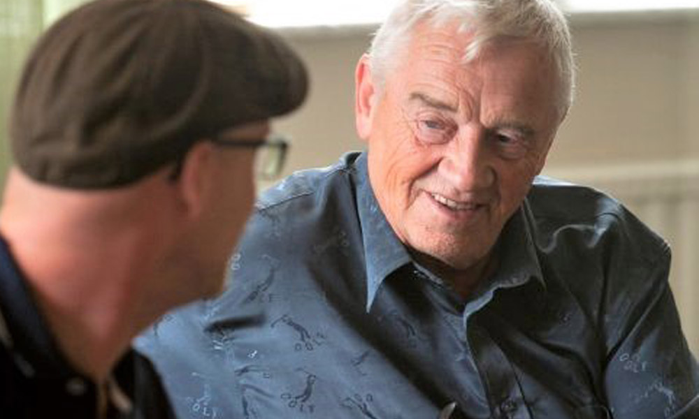 Två män samtalar och ler i en avslappnad miljö. Den äldre mannen bär en blå skjorta och den yngre en keps