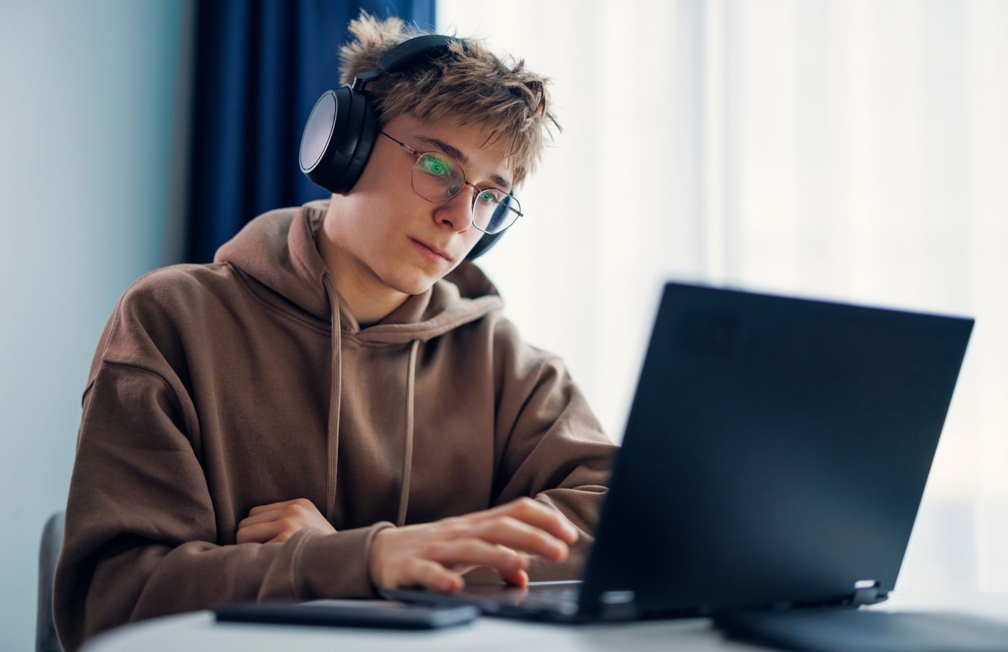 Tonårskille sitter med hörlurar och brun hoodie, arbetar vid laptop
